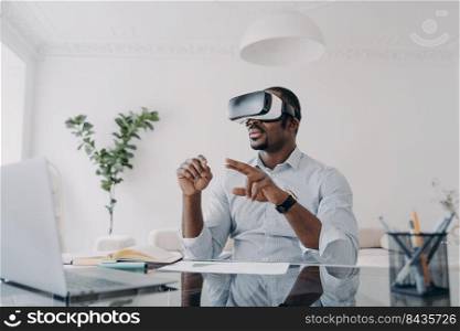 African american businessman using virtual reality glasses sitting at laptop at workplace in office. Modern black guy businessperson wearing VR goggles. Augmented Reality. Future high-tech.. African american businessman use virtual reality glasses working at laptop at office desk. High-tech