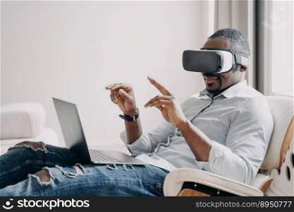 African american businessman wearing VR glasses interacting with virtual reality sitting in armchair at laptop. Black guy testing modern products in cyberspace. Hightech, future technology concept.. African american businessman in virtual reality glasses sits at laptop. Hightech, future technology