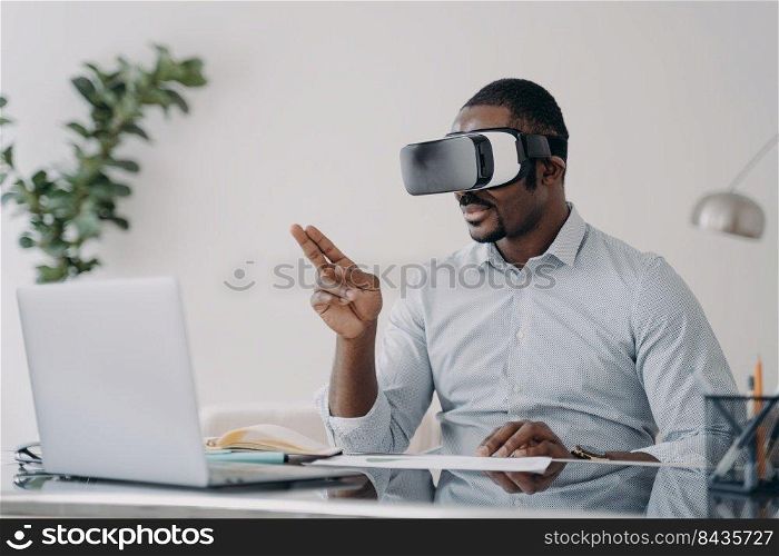 African american businessman wearing vr glasses works in augmented reality at laptop. Black young male office worker immersed in virtual reality, using innovation technology, managing business project. African businessman wearing vr glasses works at laptop manages business project in virtual reality
