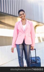 African American businesswoman commuter with roller suitcase near business office building. Black woman walking with travel bag wearing pink jacket.. Black woman walking with travel bag wearing pink jacket.