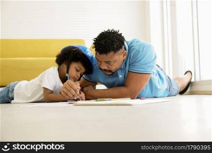 African American family father and son drawing and lying on floor at home. Young father holding his little boy hand, holding a pencil to teach drawing and painting on paper