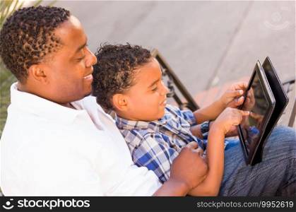 African American Father and Mixed Race Son Using Computer Tablet on Bench in Park.