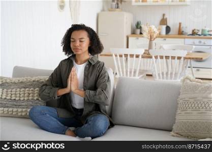 African american girl doing yoga, meditation to keep calmness, sitting in lotus pose on sofa at home. Calm young lady meditates to reduce stress, relaxing on couch. Healthy lifestyle concept.. African american girl doing yoga, meditation to keep calmness, sitting in lotus pose on sofa at home