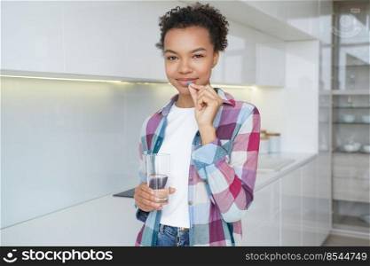 African american girl holds pill and glass of water, taking vitamins and minerals for female skin health standing at home. Vitamin deficiency prevention, healthcare, skincare concept.. African american girl holds pill, glass of water, taking vitamins, dietary supplement for wellness