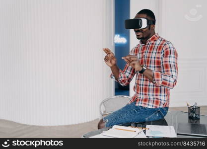 African american guy in VR glasses interacting with augmented reality, making purchases online. Black man uses virtual reality goggles pointing in the air with finger sitting at office desk. Ecommerce. African american guy in VR glasses interacts with augmented virtual reality for shopping, ecommerce