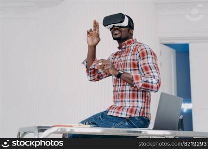 African american guy software developer wearing virtual reality glasses, creates app, working in cyberspace, sitting on office desk. Modern guy in vr goggles interacts with virtual object on workplace. African american guy software developer in virtual reality glasses creates app, sits on office desk