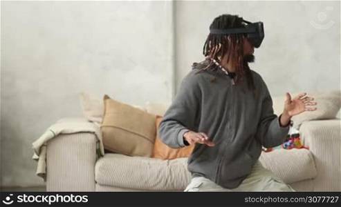 African american hipster with dreadlocks experiencing games with virtual reality goggles in modern apartment. Headset VR device playing with smartphone. Man sitting on the floor near couch, turning his head and making moves with his hands.
