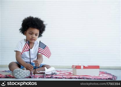 African American little boy holding small USA flags sitting on us blanket on white background, celebrating 4th July - Independence Day