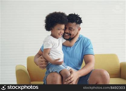 African American little son sitting on his father's lap on sofa at home. Young dad with beard hug and smile at his boy