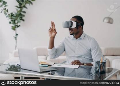 African american male architect wearing virtual reality glasses working on project in VR on laptop, sitting at office desk. Modern black businessman works in cyberspace using high tech gadget.. African american man architect in virtual reality glasses works on project in VR on laptop in office