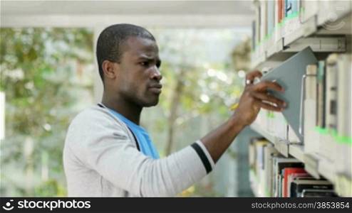 African american male college student taking book from shelf in library and looking at camera
