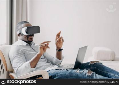 African american male software developer in virtual reality glasses tests new app, working on laptop, sitting in chair. Modern businessman touching objects in cyberspace, uses high tech for business.. African american male software developer in virtual reality glasses tests new app, working on laptop
