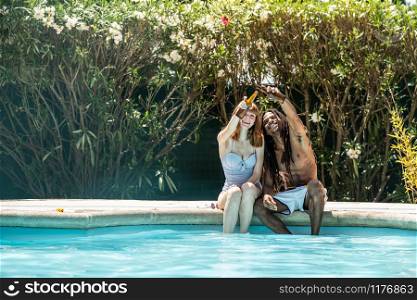 African-American man and white woman toasting with beer bottles on the edge of a pool.