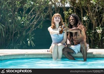 African-American man and white woman toasting with beer bottles on the edge of a pool.