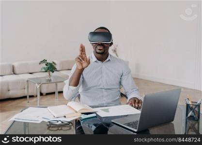 African american man businessperson uses virtual reality glasses with augmented reality technology sitting at laptop at office desk. Modern black guy working on project in cyberspace wears VR goggles. African american businessman using virtual reality glasses working at laptop. Hightech, e-business