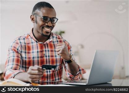 African american man holding credit card uses online banking service at laptop, makes yes gesture. Modern black guy happy with successful money transfer, cash back. E-banking, e-commerce.. African american man holding credit card uses online banking service at laptop, makes yes gesture