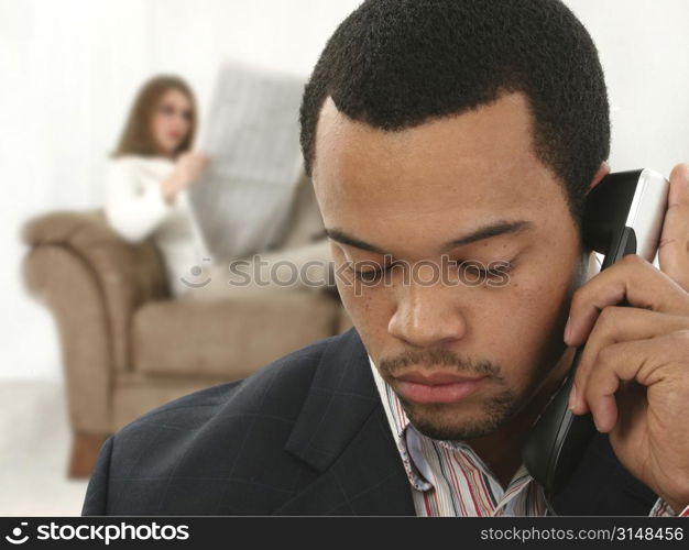 African American man in suit on landline with serious expression. Caucasian woman on couch reading newspaper in background.