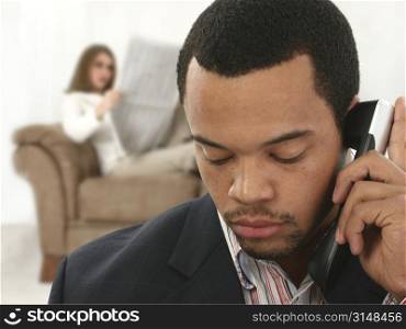 African American man in suit on landline with serious expression. Caucasian woman on couch reading newspaper in background.
