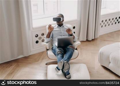 African american man in VR glasses working in augmented reality, sitting in armchair with laptop at home, pointing at virtual reality object, interacting with cyberspace, using modern high-tech device. African american businessman in VR glasses works in virtual reality, sitting in armchair with laptop
