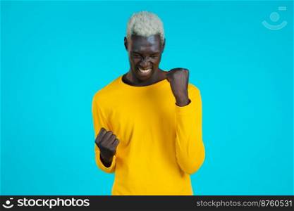African american man in yellow is very glad and happy, he shows yes gesture of victory, guy achieved result, goals. Surprised excited happy boy on blue background.. African american man in yellow is very glad and happy, he shows yes gesture of victory, guy achieved result, goals. Surprised excited happy boy on blue background