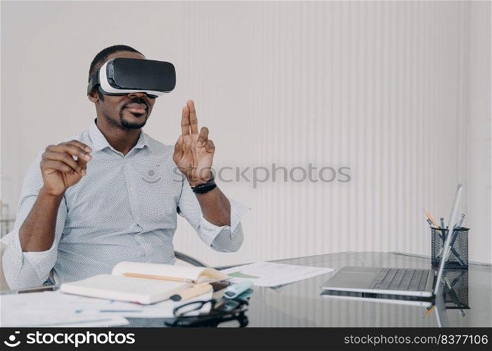 African american man IT engineer developer in VR glasses interacting with virtual objects or interface working at office desk. Modern black male businessperson using virtual reality goggles.. African american man IT engineer developer in VR glasses working with virtual reality at office desk