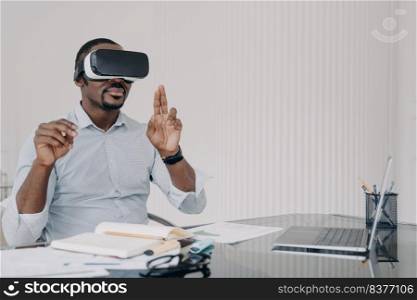 African american man IT engineer developer in VR glasses interacting with virtual objects or interface working at office desk. Modern black male businessperson using virtual reality goggles.. African american man IT engineer developer in VR glasses working with virtual reality at office desk