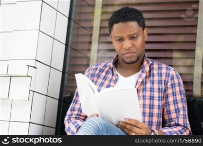 African american man relaxed and reading a book while sitting at a store window on the street.