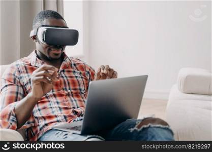 African american man software developer wearing virtual reality glasses working in cyberspace, creating app at laptop. Modern black guy programmer freelancer in VR goggles creates game application.. African american man software developer wearing virtual reality glasses works, creates app at laptop