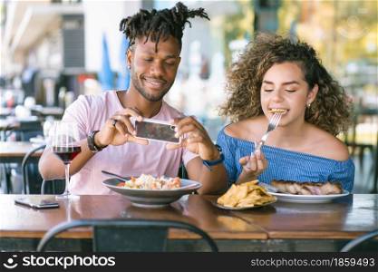 African American man taking photos to the food while having lunch with his girlfriend at a restaurant.
