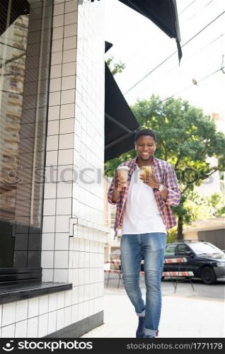 African american man using his mobile phone and holding a cup of coffee while walking outdoors on the street.