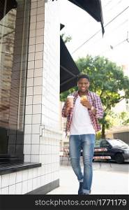 African american man using his mobile phone and holding a cup of coffee while walking outdoors on the street.