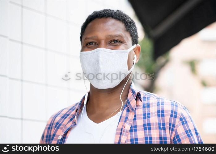 African american man wearing a face mask while listening music with earphones outdoors on the street. New normal lifestyle concept.