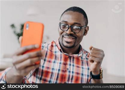 African american man wearing glasses holding smartphone having video call conversation, gesturing, takes selfie. Happy black guy looking at mobile phone smiling reading good news message.. Happy african american man wearing glasses holding smartphone smiling reading good news message