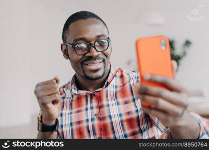 African american man wearing glasses holding smartphone having video call conversation, gesturing, takes selfie. Happy black guy looking at mobile phone smiling reading good news message.. Happy african american man wearing glasses holding smartphone smiling reading good news message