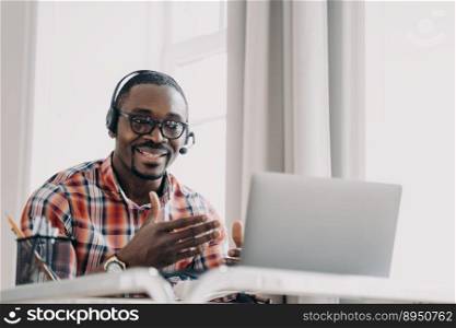 African american man wearing headset communicates with client by video call at laptop sitting at desk in home office. Friendly young black male hotline specialist helps clients remotely in workplace. African american man wearing headset communicates with client by video call at laptop. Remote work
