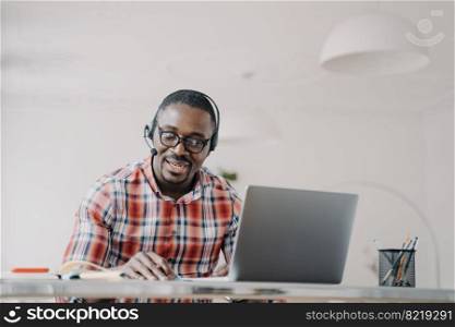 African american man wearing headset, sitting at office desk, looking at laptop screen, using app for video call. Company consultant, teacher working online. E-learning, remote communication concept.. African american man in headset works at laptop answers video call. E-learning, remote communication