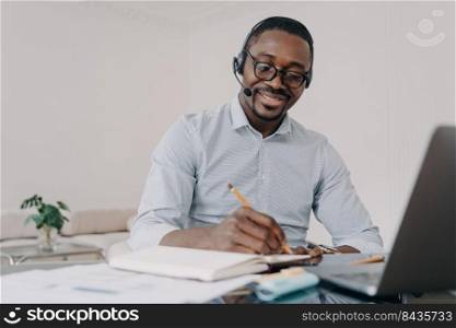 African american man wearing headset working distantly on computer at home office. Black guy student in headphones learns online at laptop, takes notes. E-learning, distance education.. African american man in headset learns online at laptop, takes notes. Elearning, distance education