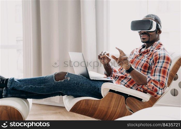 African american man wearing VR glasses making purchases in augmented reality store, play games. Modern black young guy working in cyberspace sitting in armchair with laptop. E commerce and tech. African american man in VR glasses making purchases in augmented reality store at laptop. E commerce
