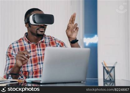 African american man wearing VR glasses working on laptop, immersed in augmented virtual reality, sitting at office desk. Black young businessman using innovation technology for work and business.. African american man wearing VR glasses working on laptop in virtual reality, sitting at office desk