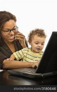 African American mid adult businesswoman at work on laptop and cell phone with toddler son on lap.