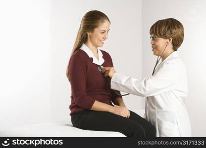 African American middle-aged female doctor examining Caucasian mid-adult female patient with stethoscope.