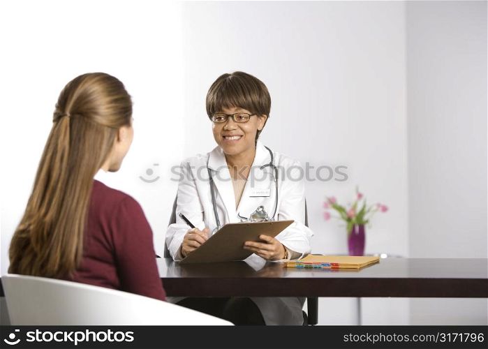 African American middle-aged female doctor sitting at desk talking to Caucasian mid-adult female patient taking notes on clipboard.