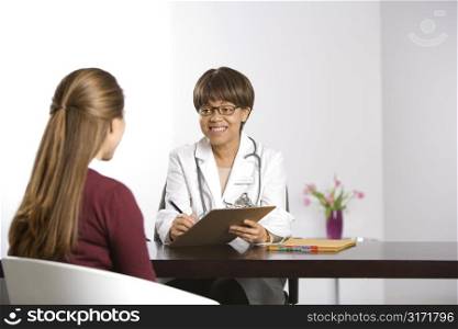 African American middle-aged female doctor sitting at desk talking to Caucasian mid-adult female patient taking notes on clipboard.