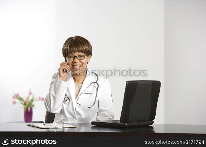 African American middle-aged female doctor sitting at desk working on laptop, talking on cell phone, smiling and looking at viewer.