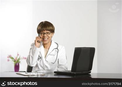 African American middle-aged female doctor sitting at desk working on laptop, talking on cell phone, smiling and looking at viewer.