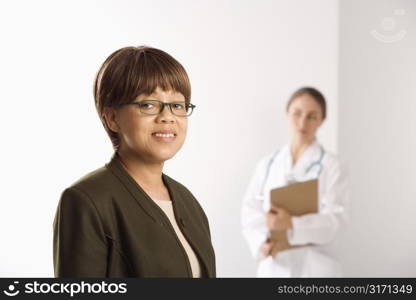 African American middle-aged female patient woman smiling looking at viewer with Caucasian mid-adult female doctor standing in background.