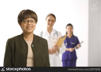 African American middle-aged woman smiling and looking at viewer with Caucasian mid-adult female doctor and Asian Chinese mid-adult female physician&acute;s assistant standing in background.