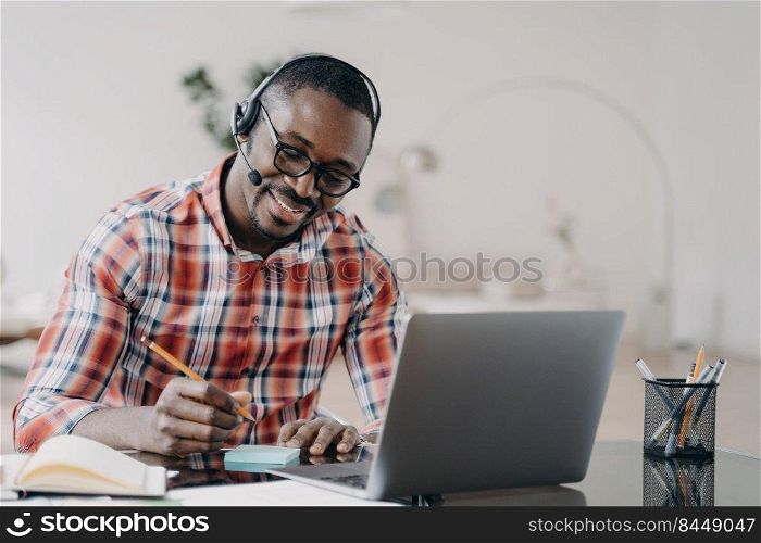 African american student young man getting remote education at laptop. Happy black male wearing headset listening lecture, taking notes while online lesson by video conference. Distance learning . African american student in headset learning at laptop online, makes notes. Distance education