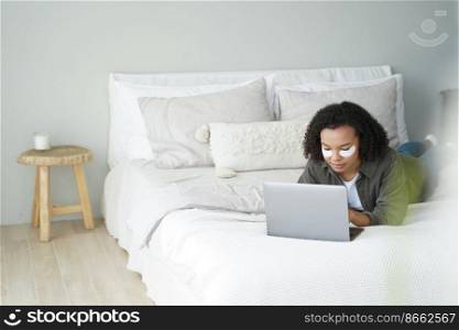 African american teen girl with under eye patches working on laptop lying on bed at home during daily skincare routine. Biracial young lady shopping online on computer while morning beauty procedure.. Biracial teen girl with under eye patches shopping online at laptop in bedroom while beauty routine