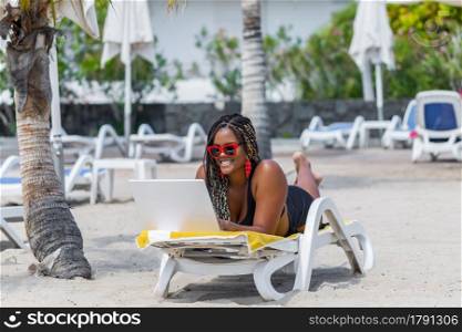 African American woman on the beach lying on a deck chair using a laptop. Cheerful adult woman enjoying a summer day at the beach.. African American woman on the beach lying on a deck chair using a laptop